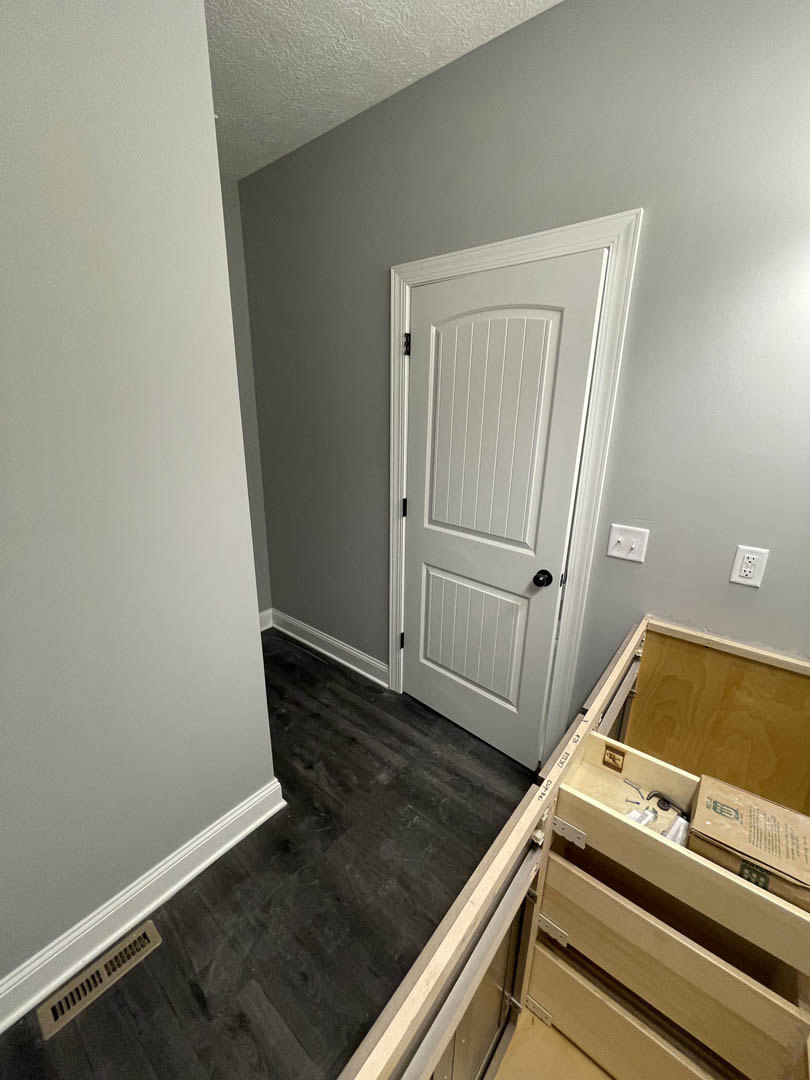 White paneled door with black knob, white light switch, black hardwood floor with white baseboard trim, wall vent, and brown box with black text against plaster walls.