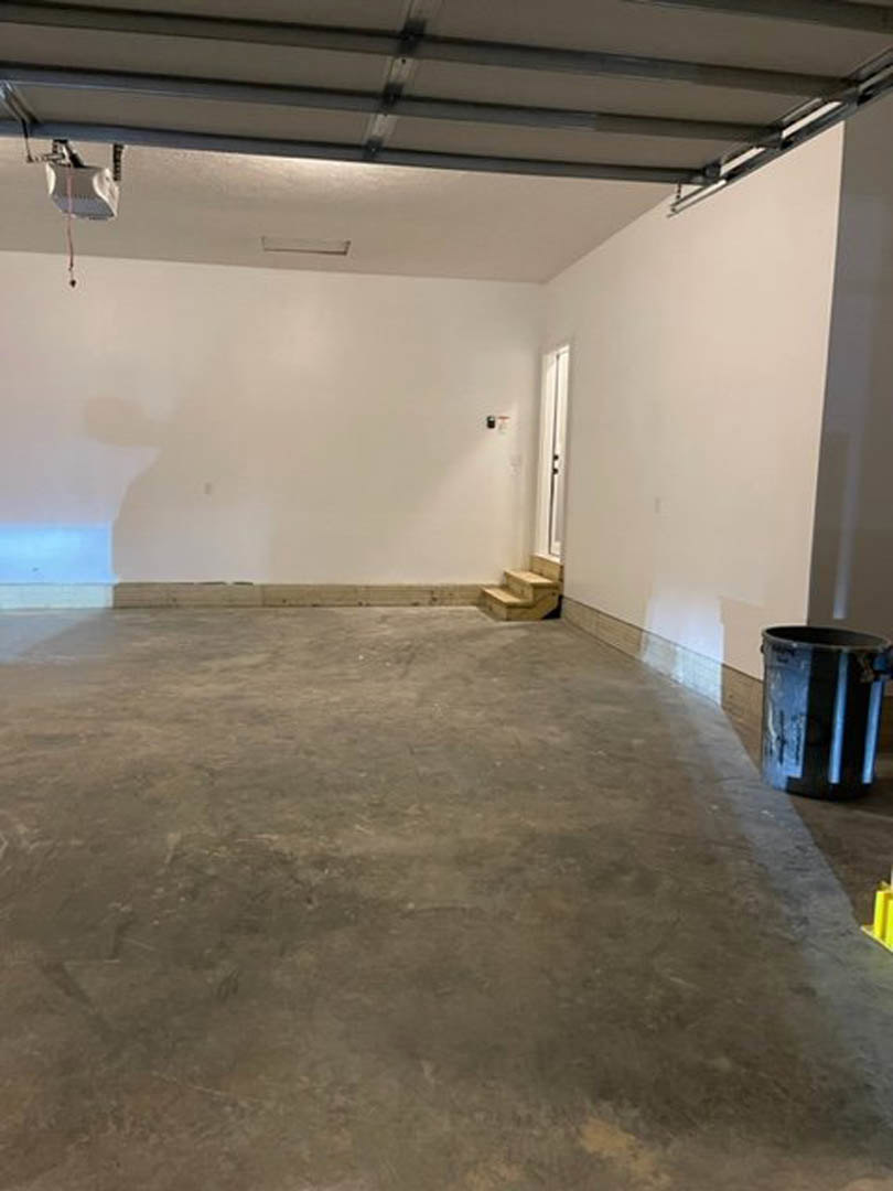 Garage interior with concrete floor, white plaster walls, blue trash can near a closed door, metal ceiling with mounted light, staircase in the corner