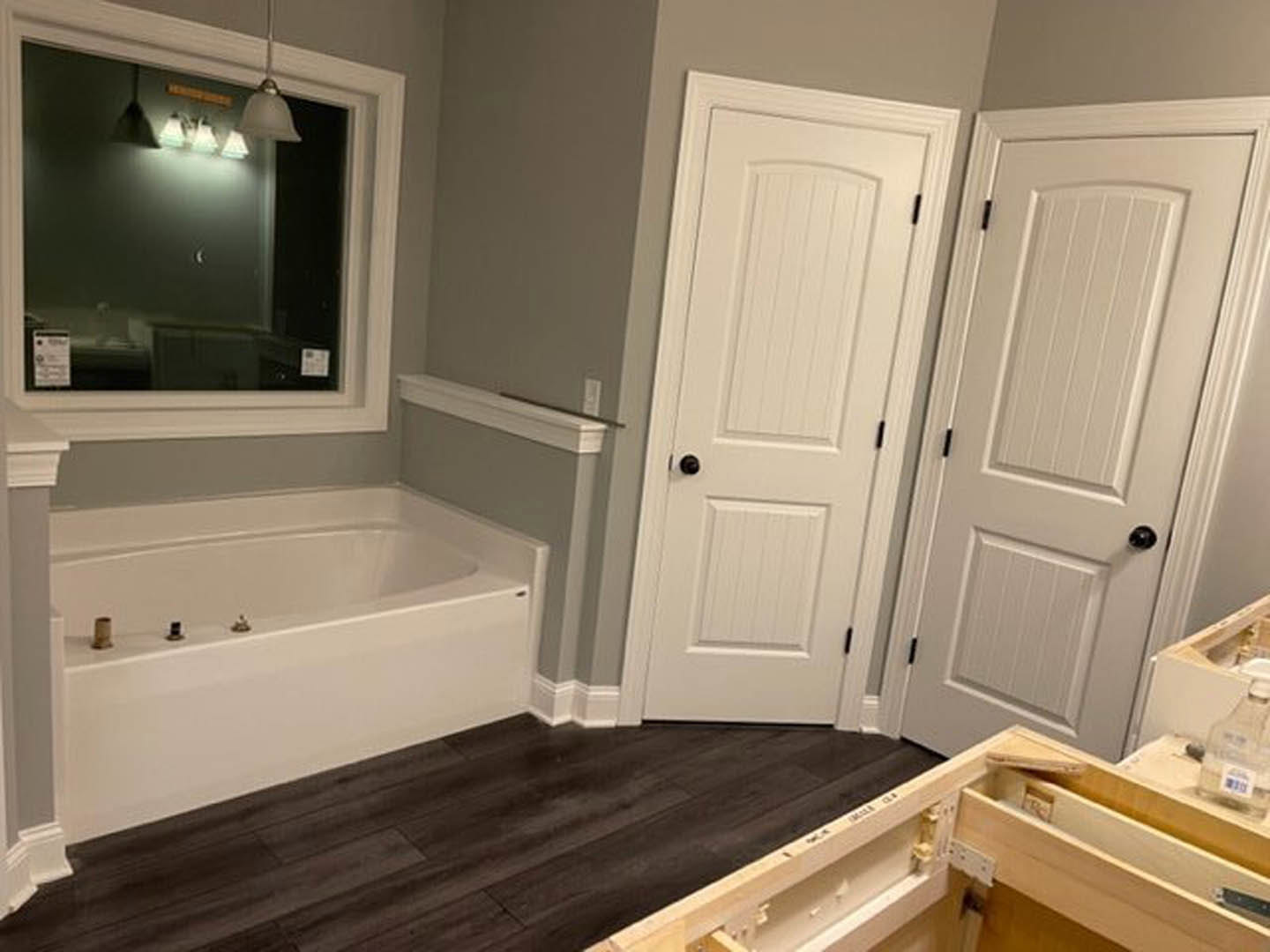 Bathroom with freestanding bathtub, two white doors with black knobs, dark wood flooring, white walls, window letting in natural light, and modern plumbing fixtures
