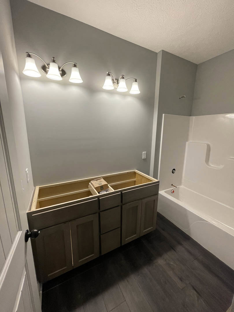 Freestanding white bathtub beside dark wood cabinets, light gray tile floor, white quartz countertop, three-bulb wall sconce with frosted glass shades