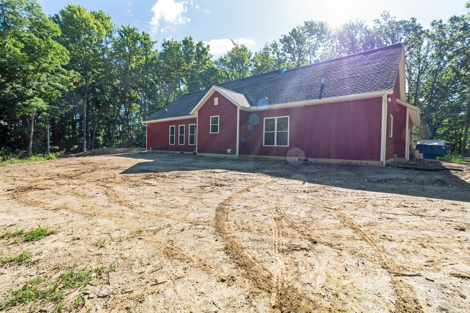 Single-story home with white-framed windows, dirt yard featuring tire tracks, scattered trees in the background, blue fence partially visible, cloudy sky overhead