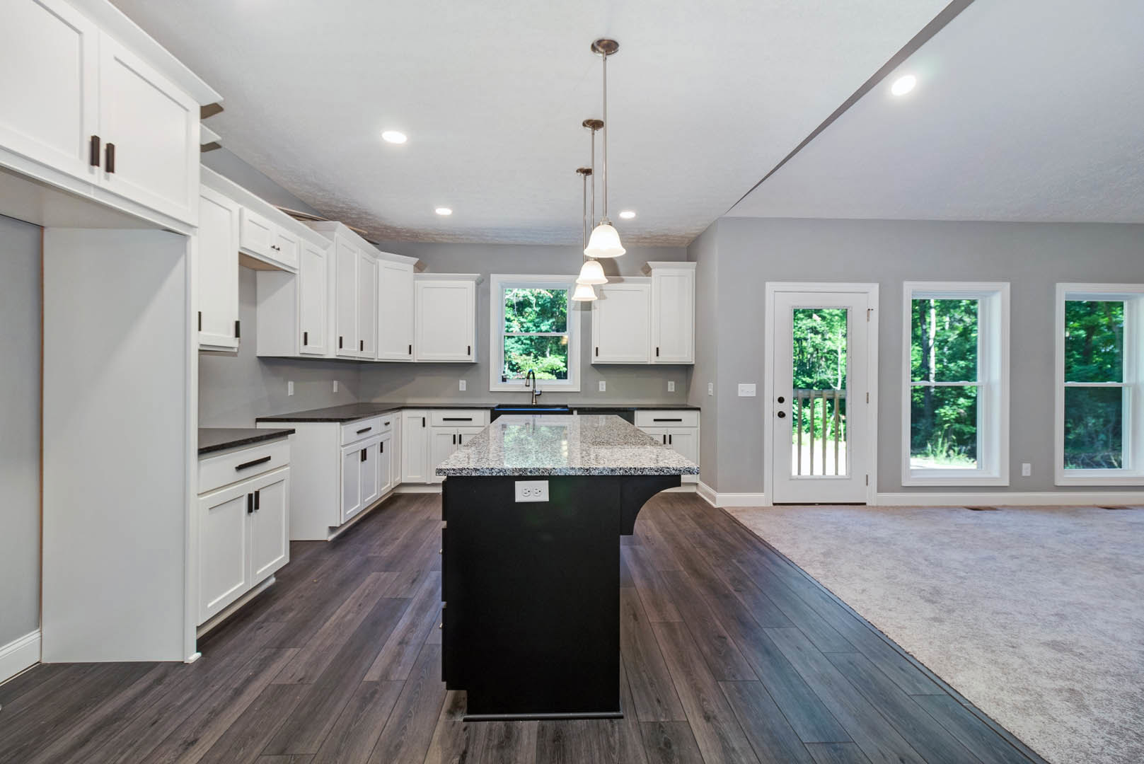 White kitchen cabinets, black island with marble countertop, stainless steel sink, bell-shaped pendant light, window overlooking trees, white door with glass panel and railing