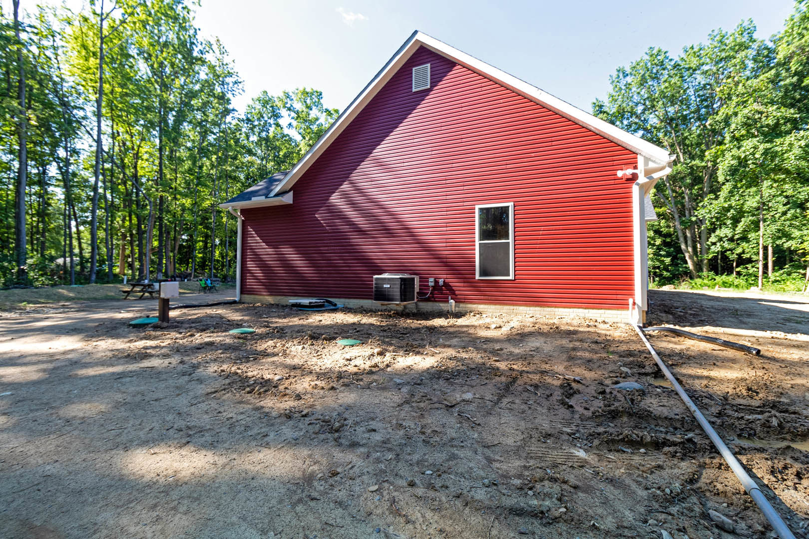 Red siding house with white metal roof, screened side window, black utility box with white label, bare dirt yard, surrounding woodland trees, partially visible white box in