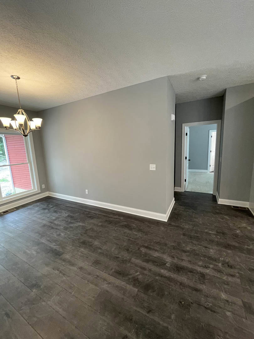 Dark wood floor in a spacious room with a white-framed window, open door, plaster walls, and a ceiling-mounted chandelier with multiple lights.