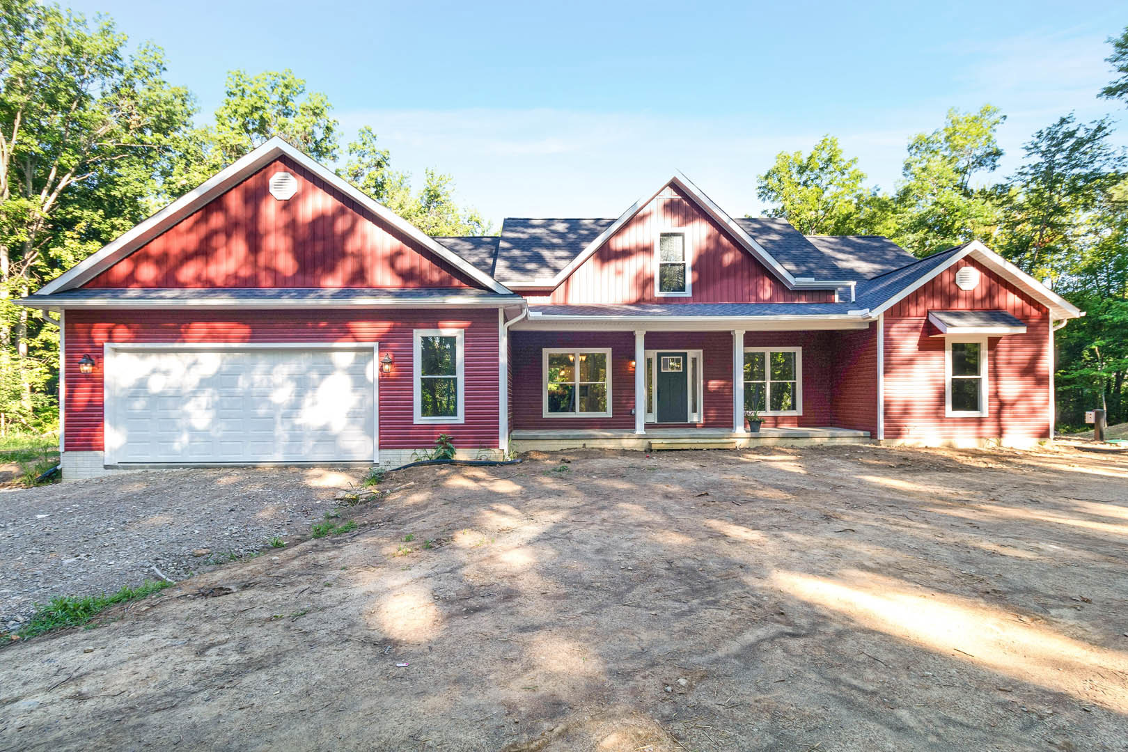 Red house with white trim and white garage door, red roof, white-framed windows, dirt yard, trees in background