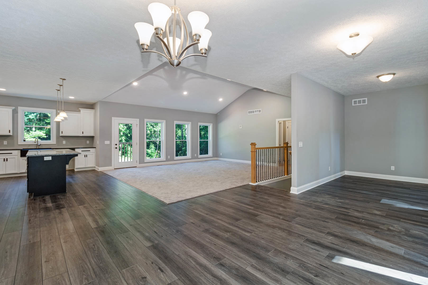 Spacious room with polished hardwood floors, ornate chandelier hanging from white ceiling, black kitchen island, white rug, and decorative railing
