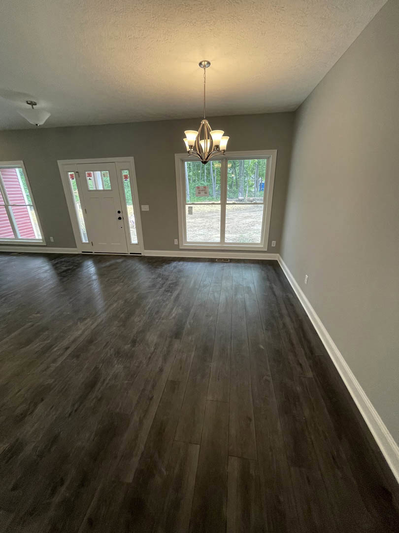 Open living space with wide plank wood flooring, white walls, large window with red shutter, glass-paneled white door, and ornate chandelier hanging from plaster ceiling