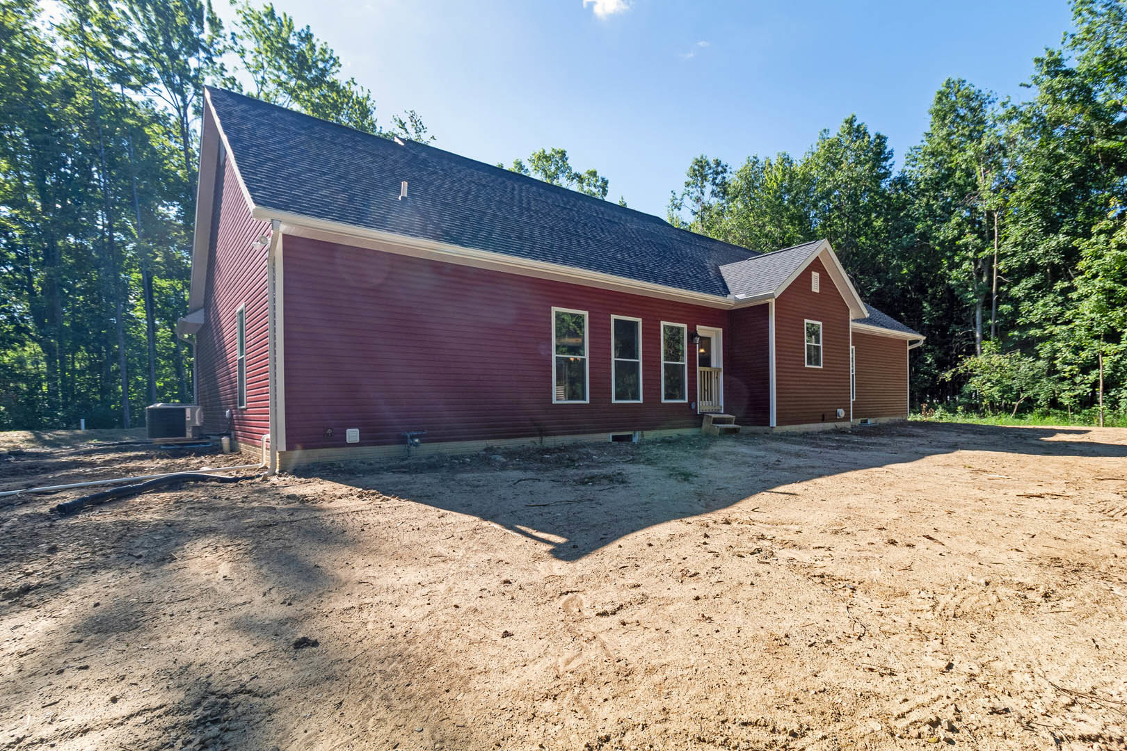 Red house with white-framed windows, covered porch, and gabled roof, surrounded by dirt lot and mature trees under partly cloudy sky