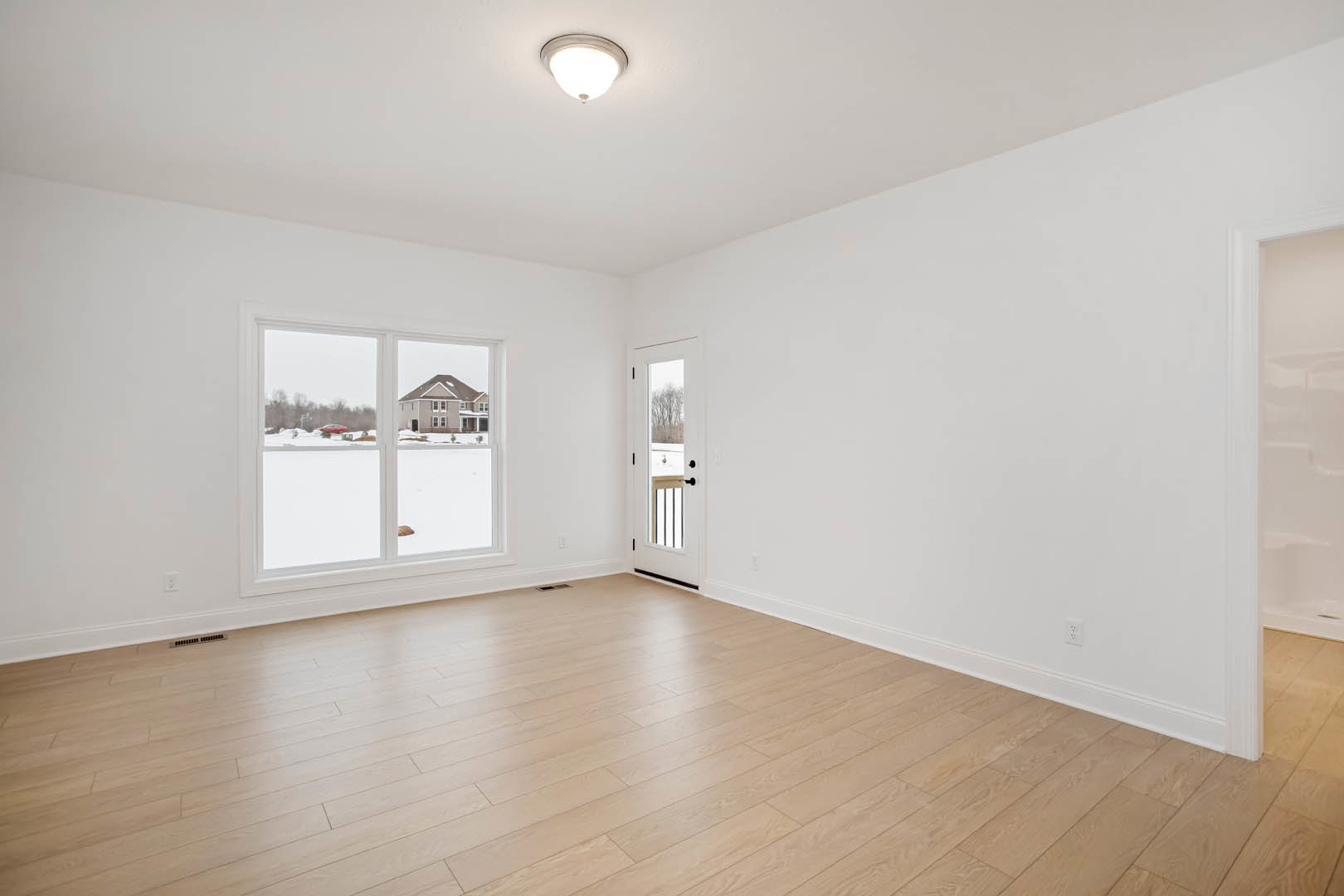 Hardwood floor room with white plaster walls, ceiling light fixture, single door with glass window, large window showing neighboring house and roof.
