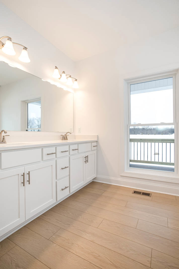 Bathroom featuring a large window with snowy view and railing, expansive mirror above white cabinetry, wooden floor with built-in drawers, row of wall-mounted lights, and tiled