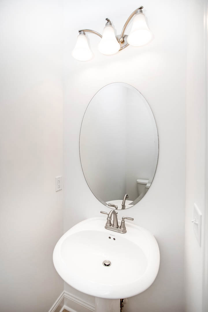 White bathroom with round wall mirror above rectangular sink, chrome faucet, and modern light fixture; partial view of toilet and neutral finishes.