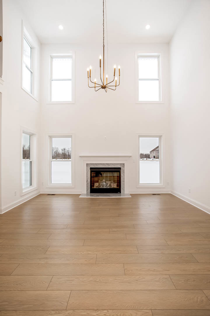 Living room with hardwood floors, plaster walls, stone fireplace, candle-style chandelier, large windows overlooking trees, and a person seated in an upholstered chair.