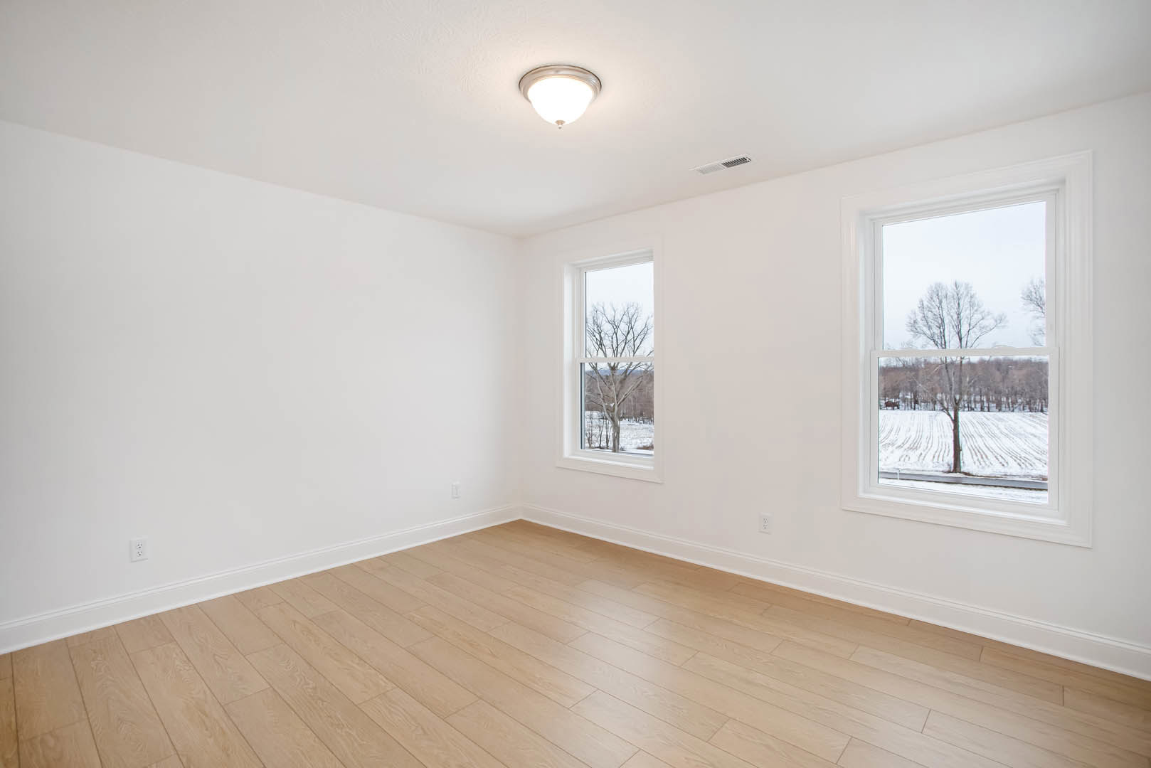 Wide-plank wood flooring in a bright room with large windows overlooking snowy trees, white plaster walls, and a modern ceiling light fixture