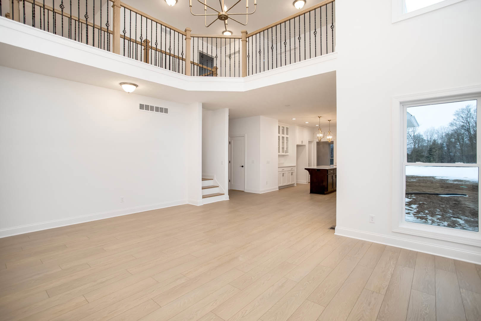 Double-height staircase with white balusters and wood treads, wood flooring throughout, kitchen island with white countertop, large windows framing tree views, white door with