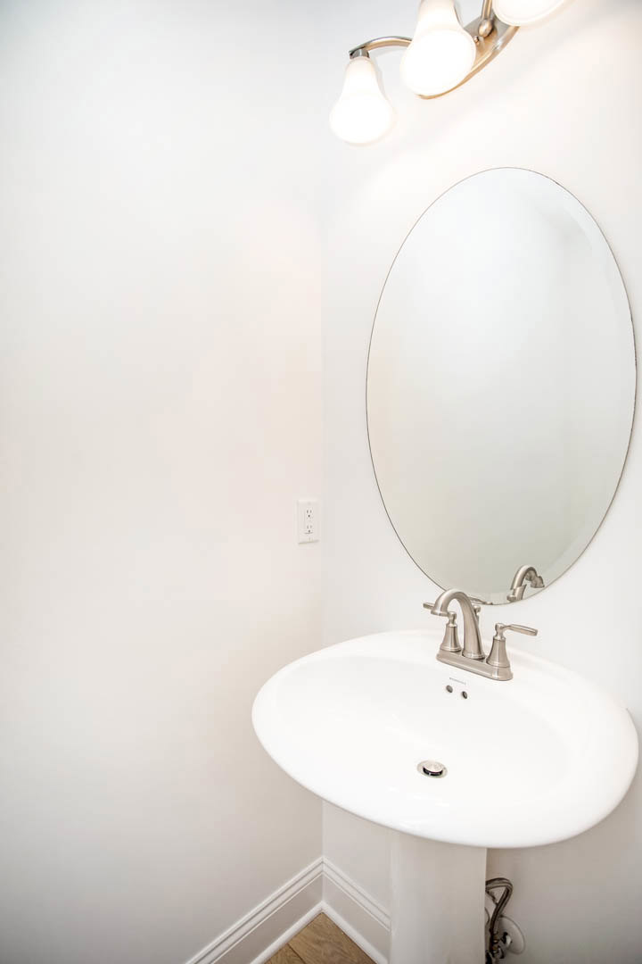 White bathroom sink with chrome faucet beneath a rectangular wall mirror, light fixture above, and corner of wood floor visible.
