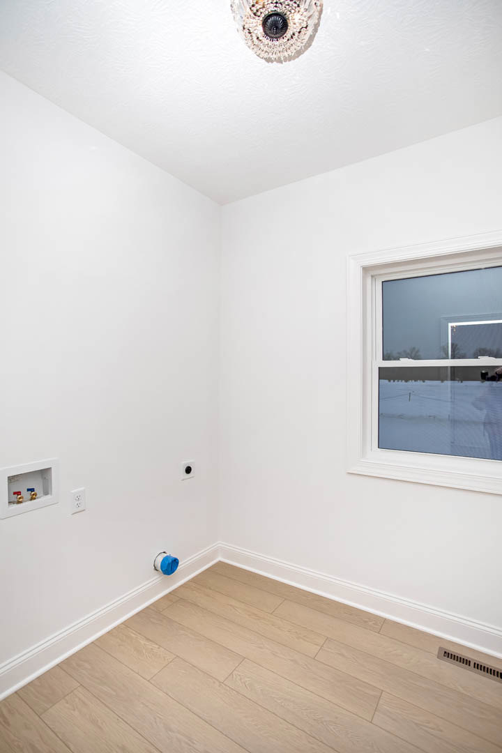 Spacious room featuring wood flooring, large window overlooking snowy landscape, modern chandelier, blue tape marking on floor, white mechanical box with valves, wall vent, and