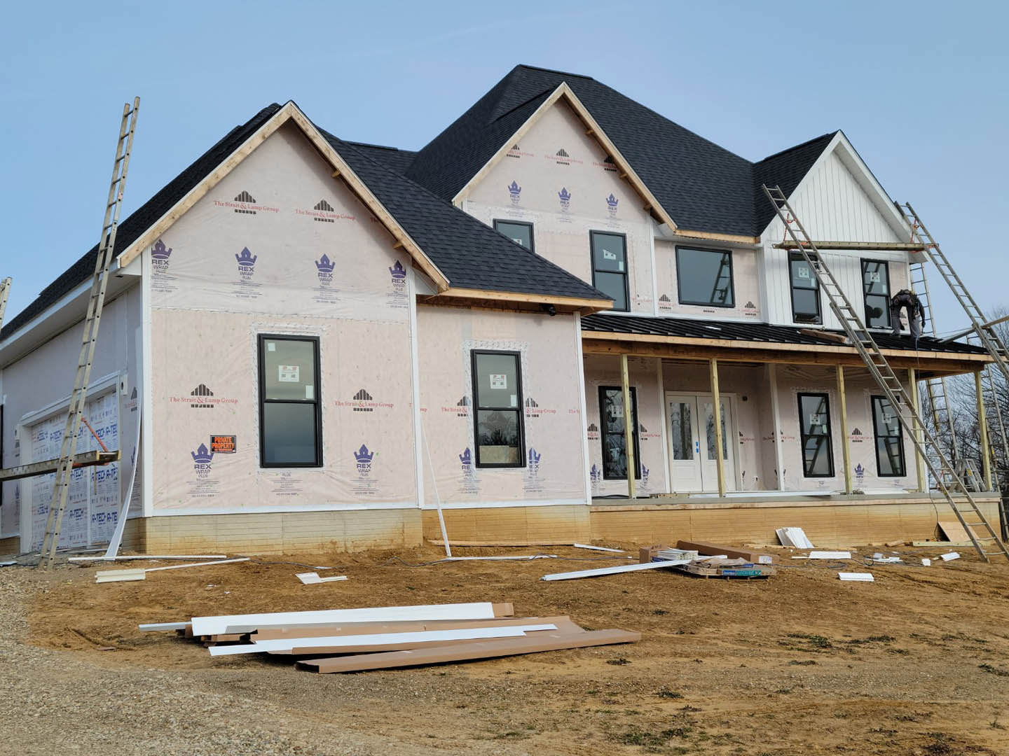 Partially finished house with black roof, exposed plywood siding, ladder leaning against exterior, and window displaying construction permit sign