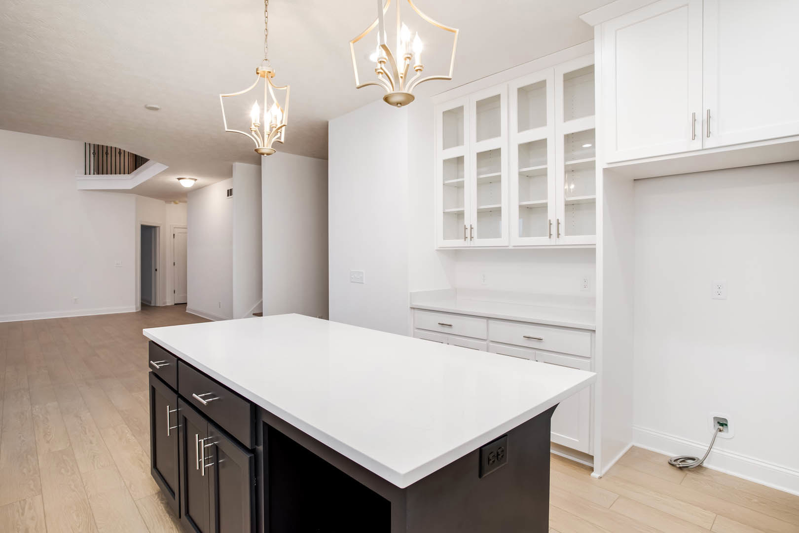 White kitchen with shaker cabinets, glass cabinet doors, white island with quartz countertop, stainless steel sink, pendant light fixtures, and light wood flooring.