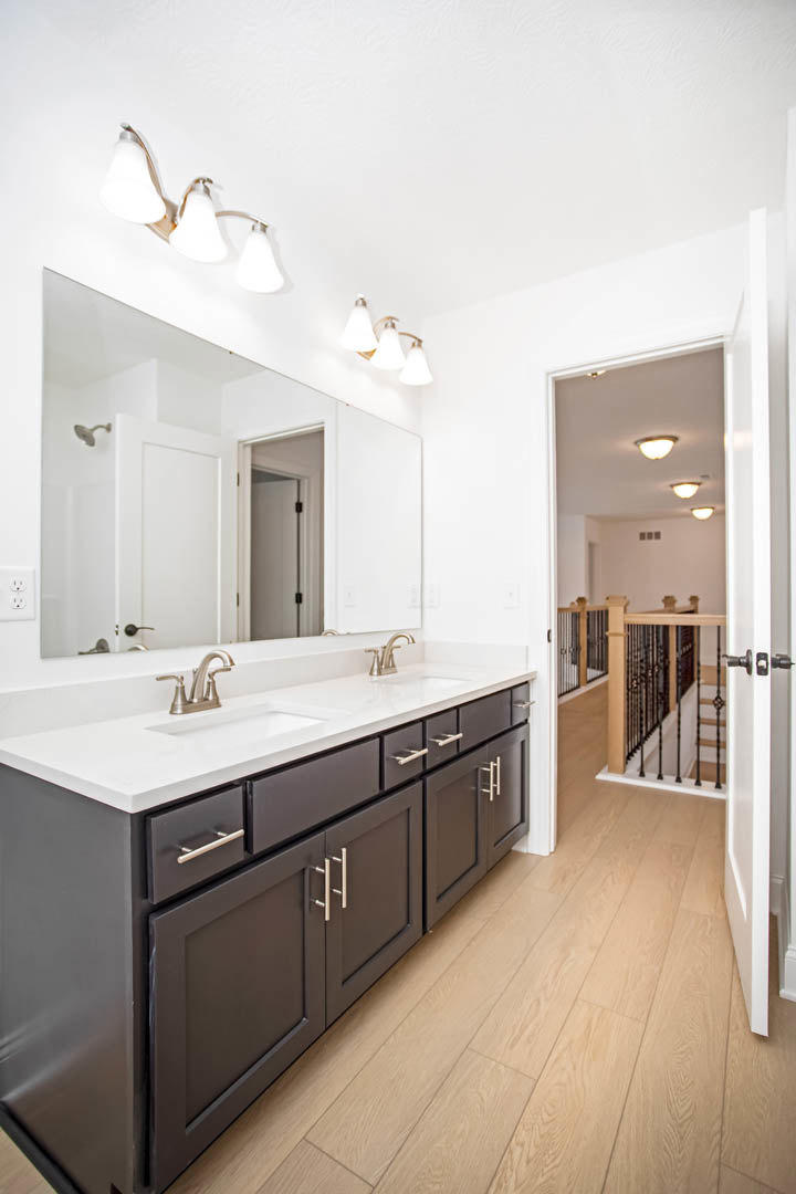Bathroom with double sinks set in black cabinetry, wide mirror above, three-light fixture mounted on wall, wooden flooring, staircase with metal railings visible in background.