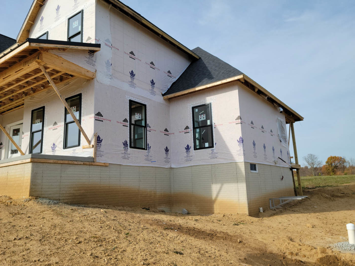 Framed house under construction with exposed wood beams, dirt hill in foreground, window featuring a temporary sign and wooden plank, white chair visible inside