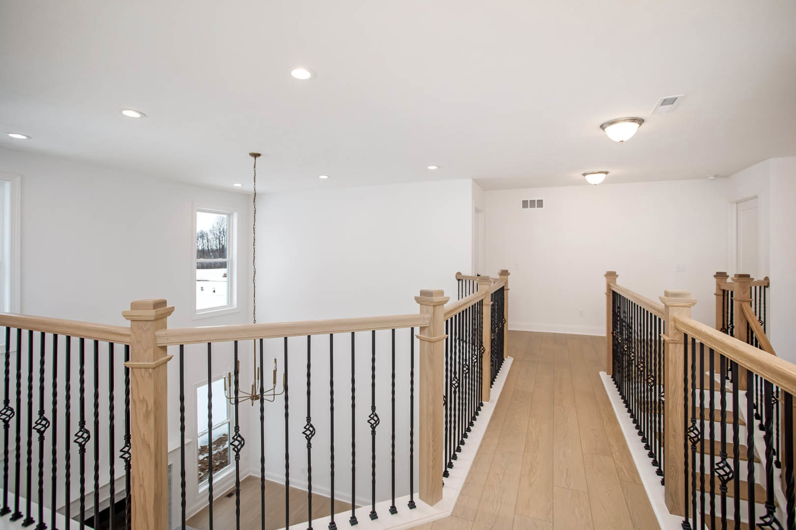 Hallway with light wood flooring, black metal railings on staircase, modern chandelier hanging from ceiling, large window overlooking snowy landscape, white square vent on wall