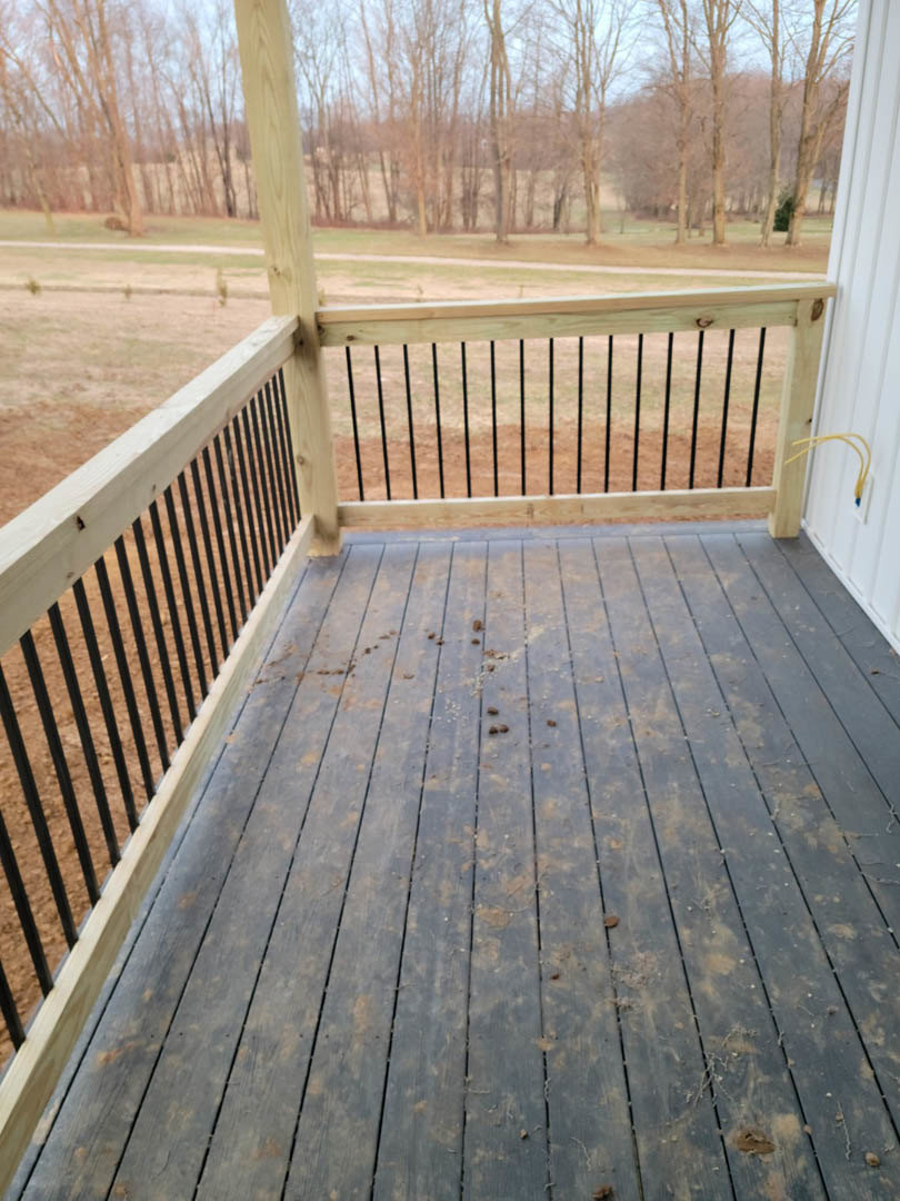 Wooden deck with metal handrail, plank flooring, and fenced perimeter, surrounded by leafy trees in the background