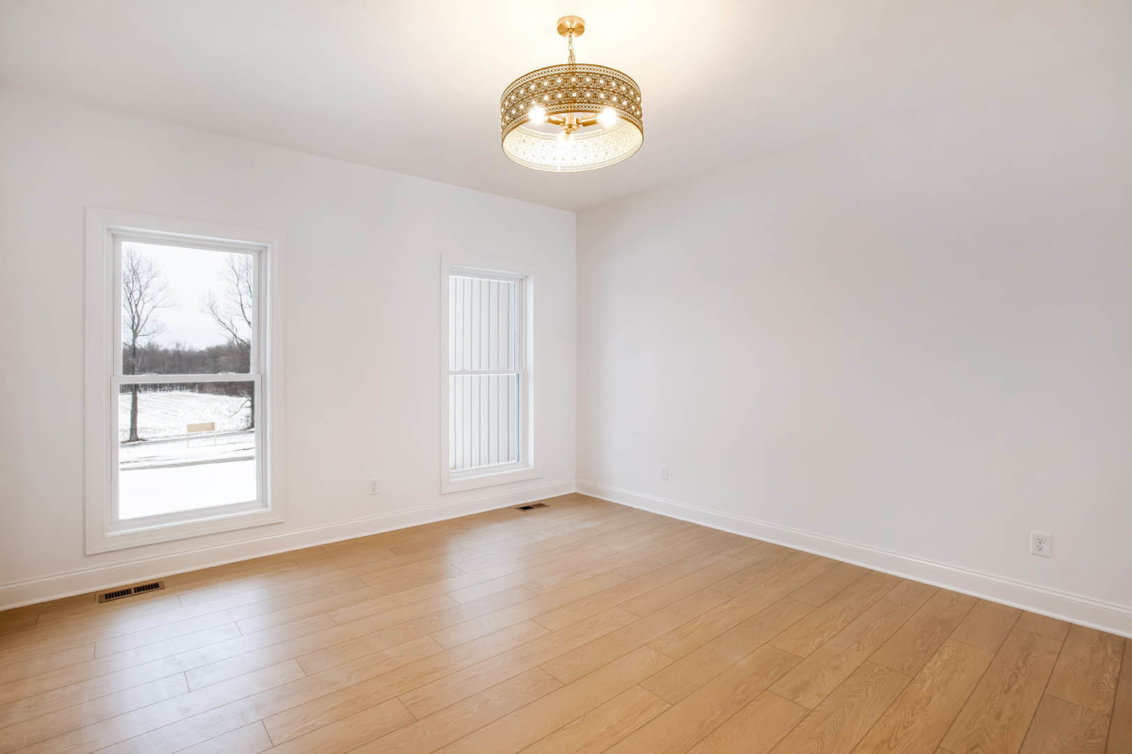 Chandelier with circular ring hangs from white ceiling above hardwood floor; large window reveals snowy field; plaster walls with crown molding.