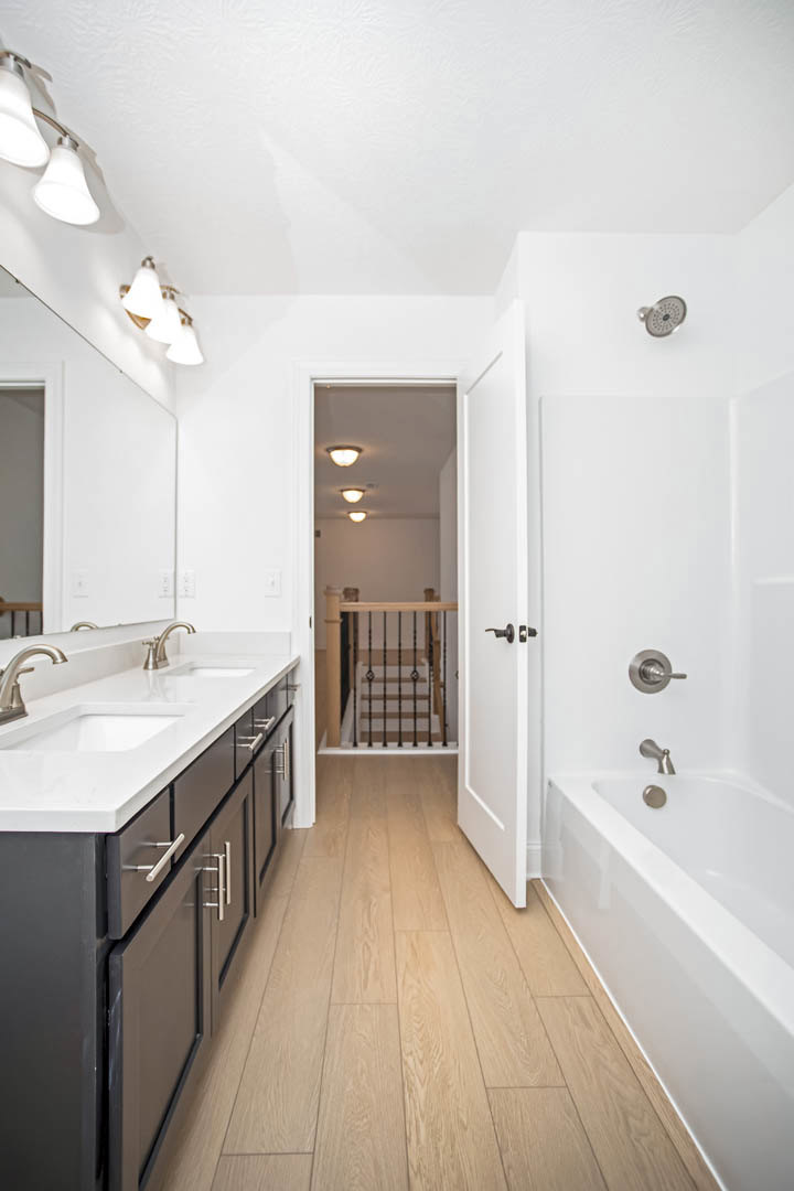 White freestanding bathtub beside wood cabinets, tile floor, undermount sink with chrome faucet, neutral wall tile