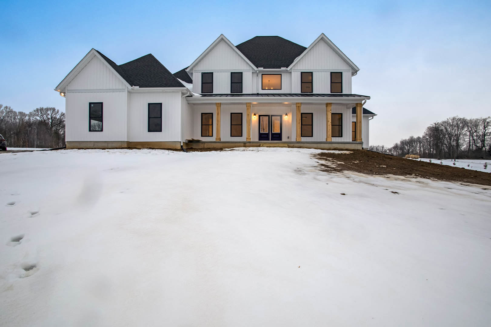 Large white house with black roof and black-trimmed windows, snow covering the ground and trees, warm light glowing from a window