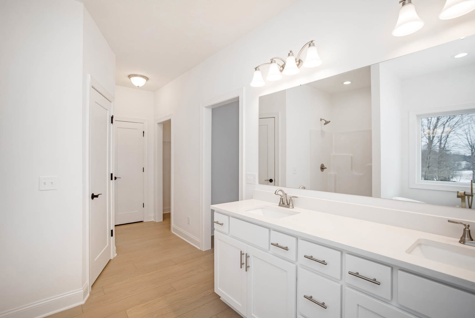 White bathroom vanity with quartz countertop, rectangular mirror above, window showing leafy trees, white door with bronze handle, ceiling-mounted light fixture, neutral tile
