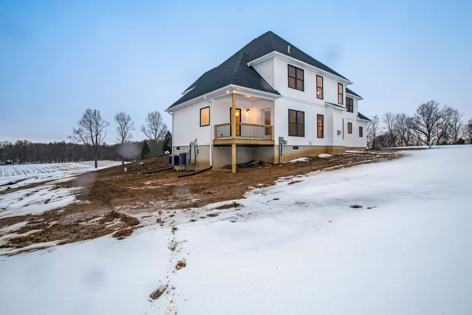 White house with black roof and deck, surrounded by snow-covered ground and trees, multiple air conditioning units visible, winter sky in background
