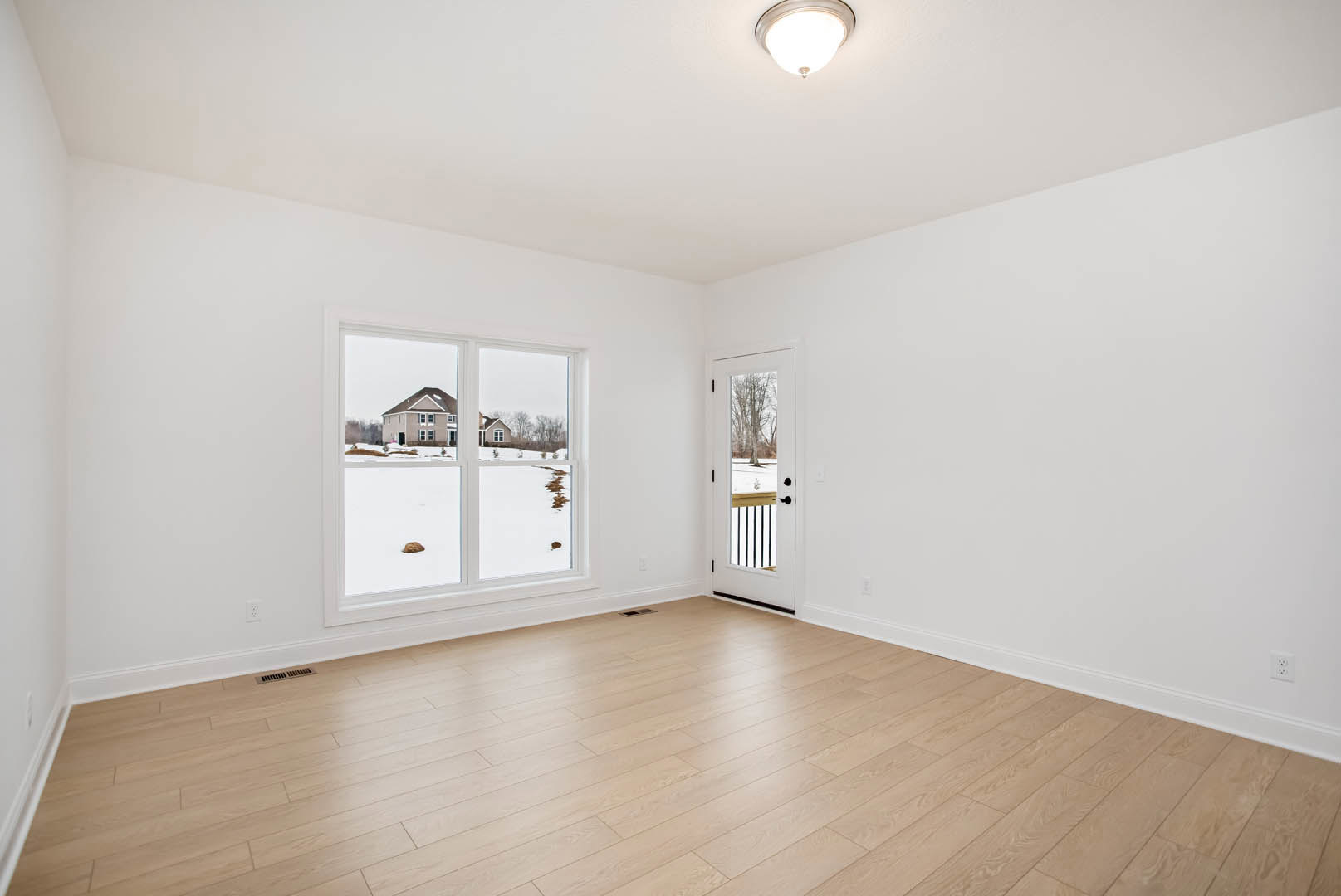Room with light wood flooring, white plaster walls, a single door opening to a snowy landscape, and a large window letting in natural light