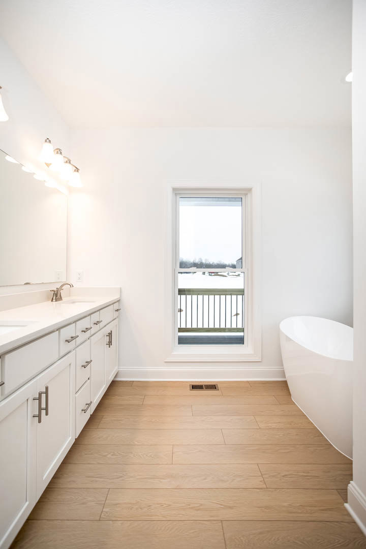 White freestanding bathtub beside a large window overlooking snowy landscape, wood floor with vent, string lights above, partial view of metal railing