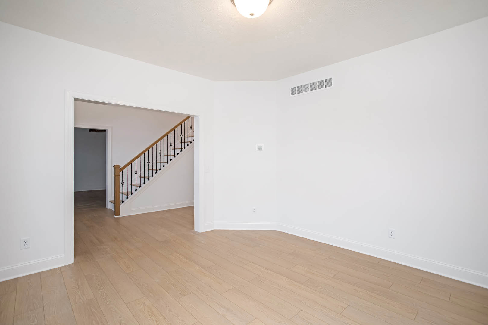 Hardwood floor room with white plaster walls, central wooden staircase, ceiling light fixture, and metal floor vent