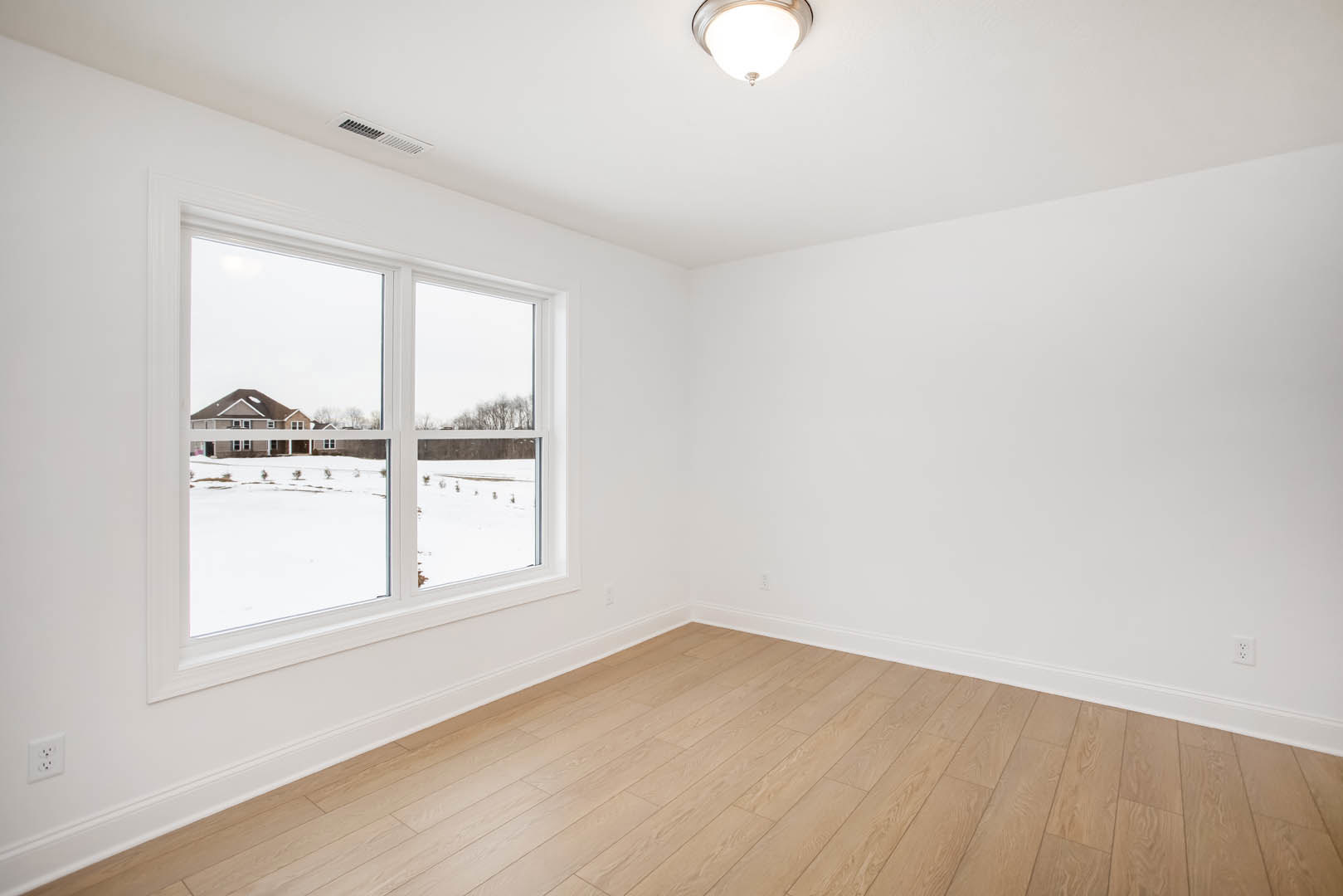 Sunlit room with wide window, white plaster walls, hardwood floor, ceiling light fixture, and white electrical outlet; exterior house visible through window.