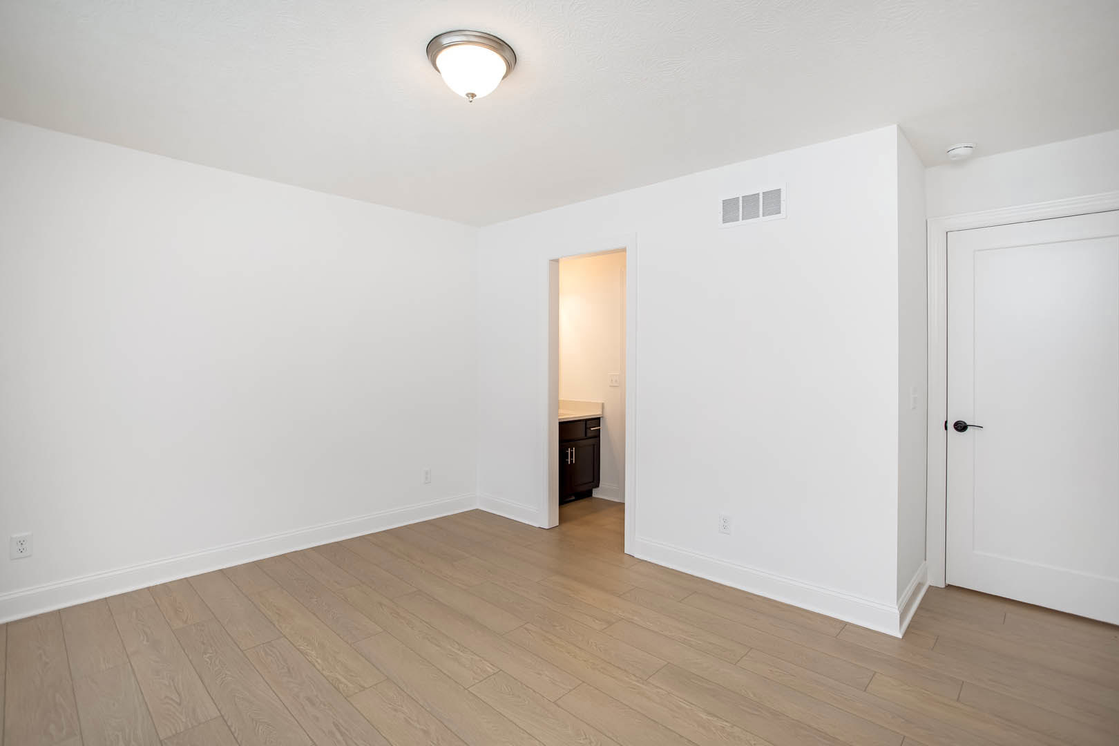 White paneled door with black handle open to a room featuring wood flooring, white baseboards, white walls with a vent, and a modern light fixture.