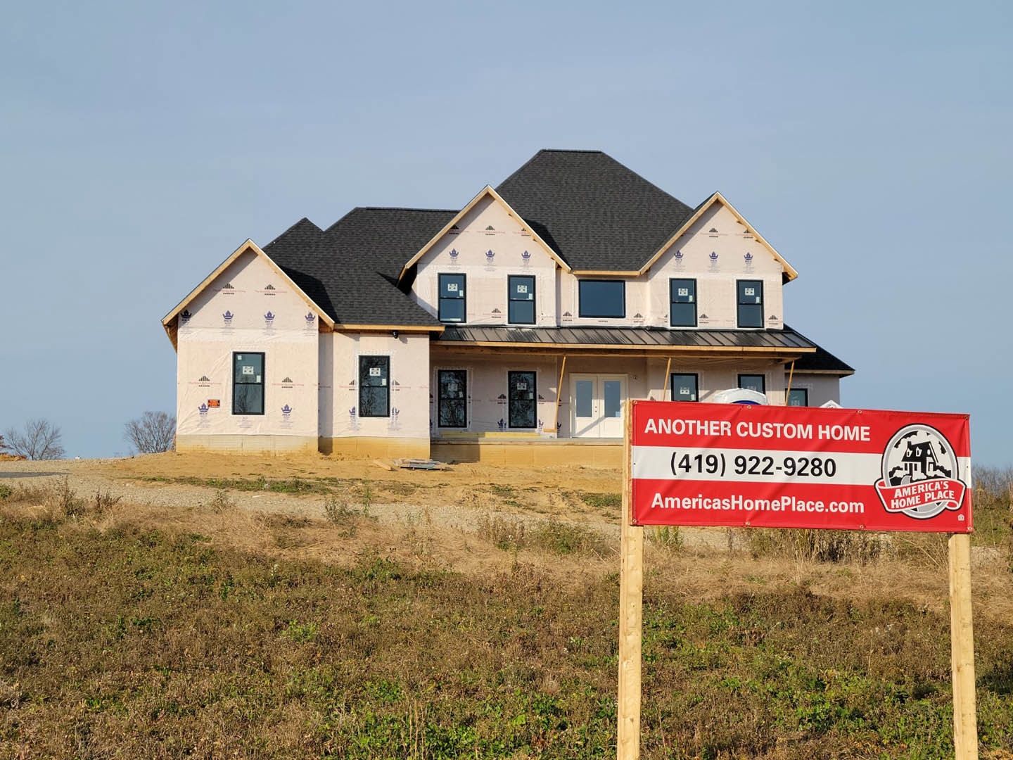 Framed house under construction with black window frames, red and white sign with black text displayed on grassy lot, blue sky overhead