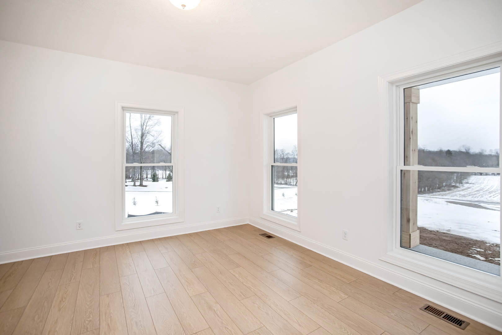 White walls and wood laminate flooring in a bright room with windows overlooking a field and trees; wall vent visible near the floor.