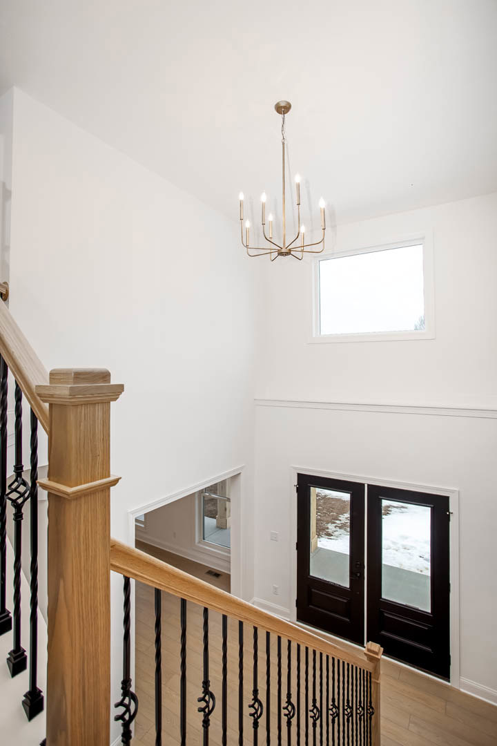 Chandelier with multiple lights hanging from ceiling, white rectangular window with white frame, double doors, staircase with wooden post and black railing, hardwood flooring