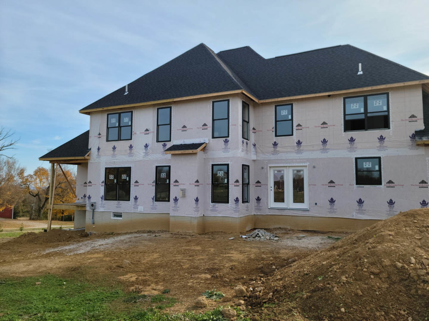 Two-story house under construction with white double doors, black-framed windows, plastic covering on exterior, dirt and grass in foreground, and a window displaying a sign.