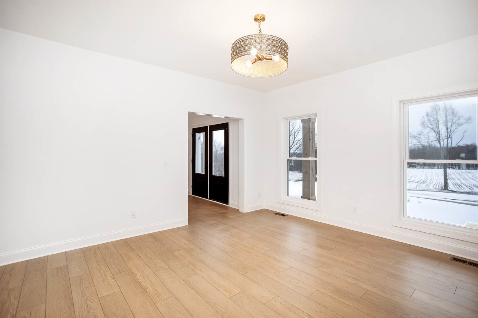 Chandelier with circular ring hanging above hardwood floors, double glass-paneled doors, large window framing snowy landscape and tree outside, plaster walls