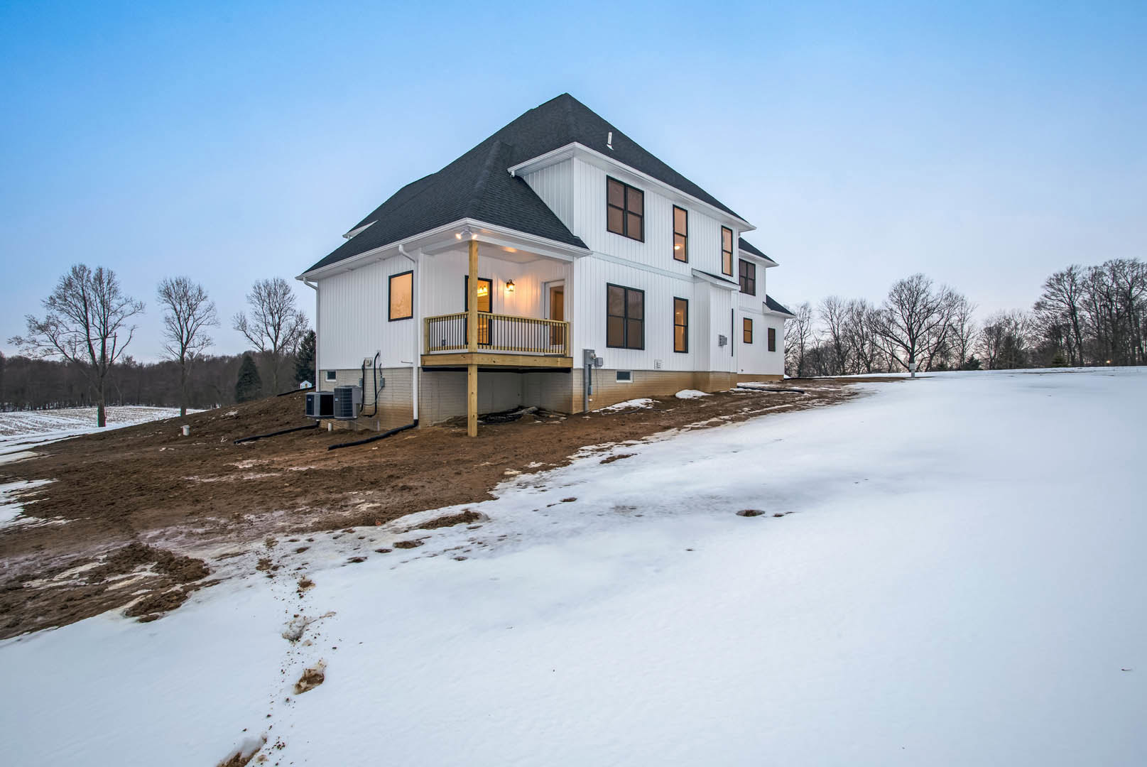White house with black roof and deck, surrounded by snow-covered ground, leafless tree, and multiple air conditioning units on the side