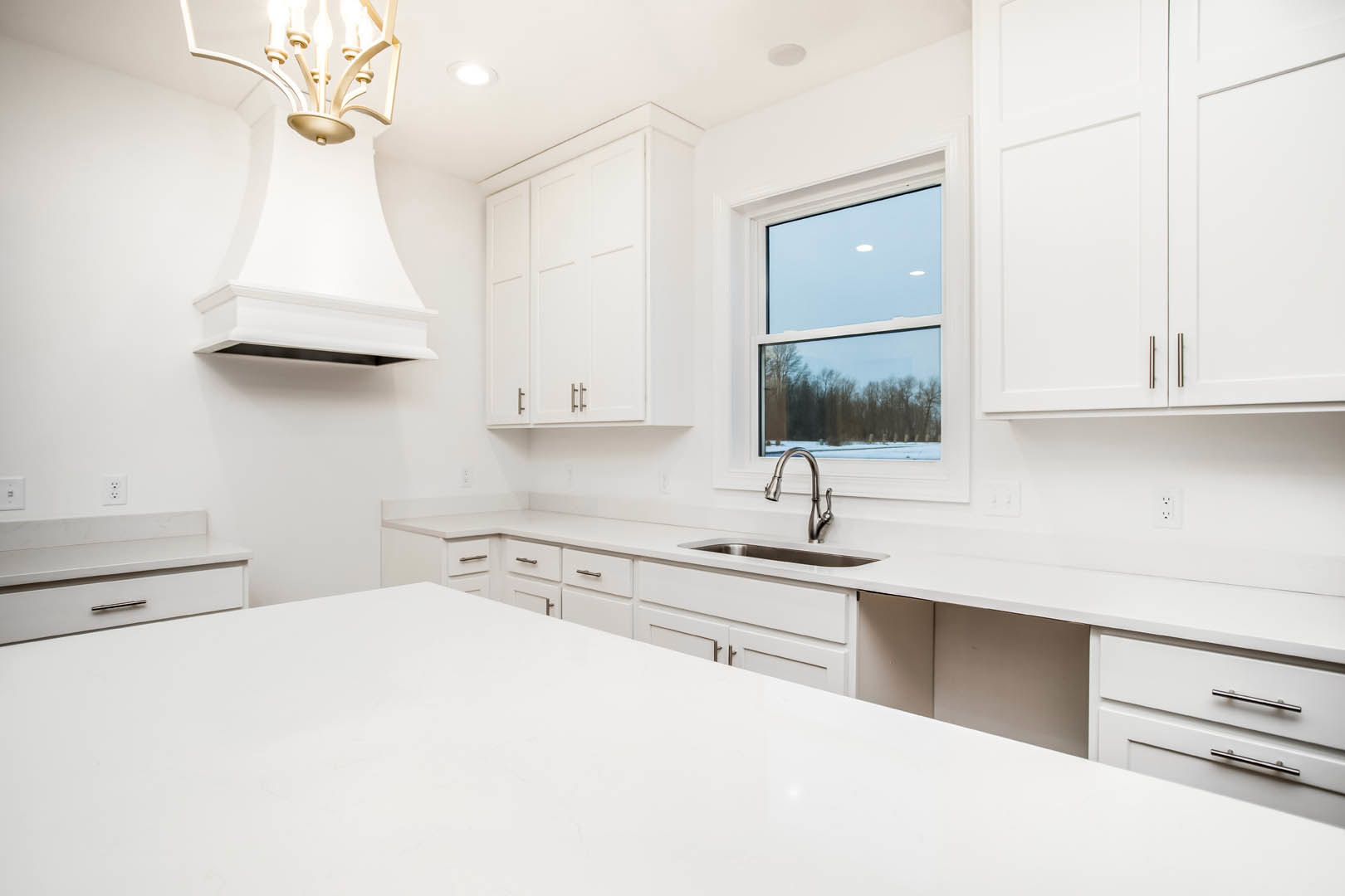 White kitchen featuring expansive white countertop, white cabinetry with three doors, stainless steel sink and tap, chandelier overhead, large window with view of green trees.
