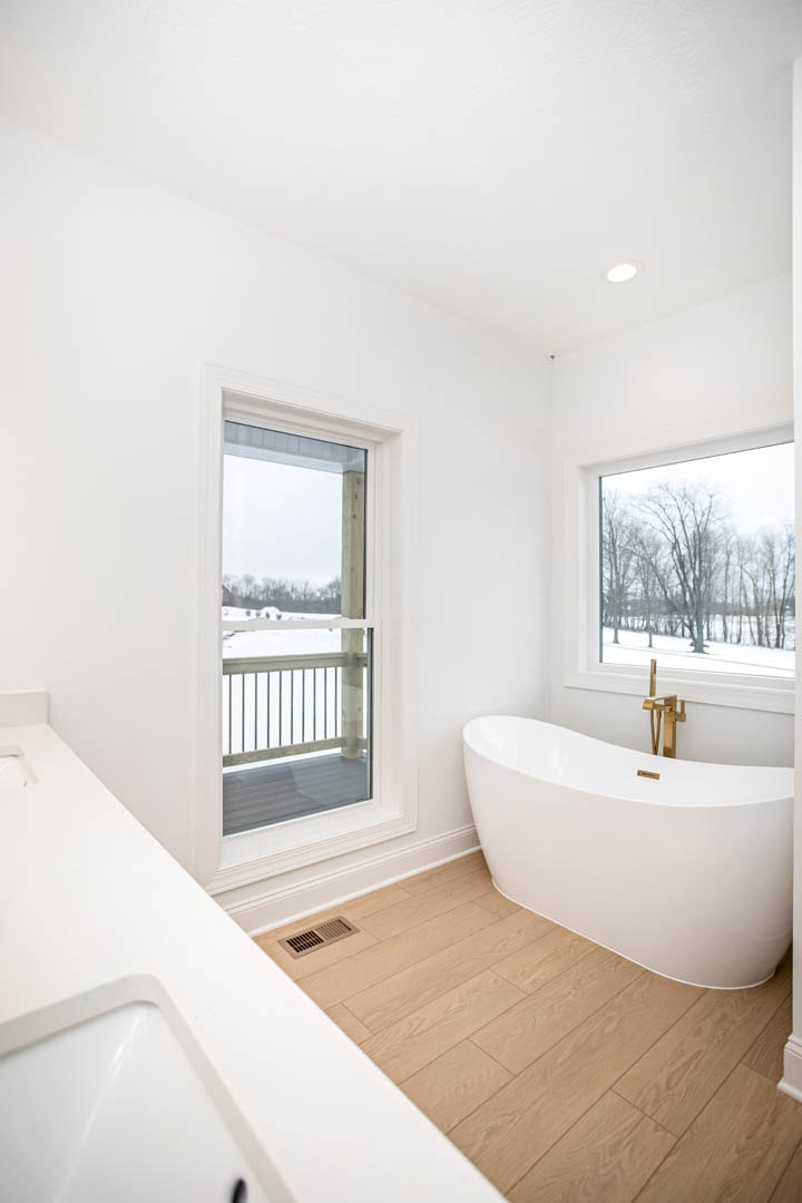 White freestanding bathtub beside large window overlooking snowy trees, wood flooring, light walls, and modern sink