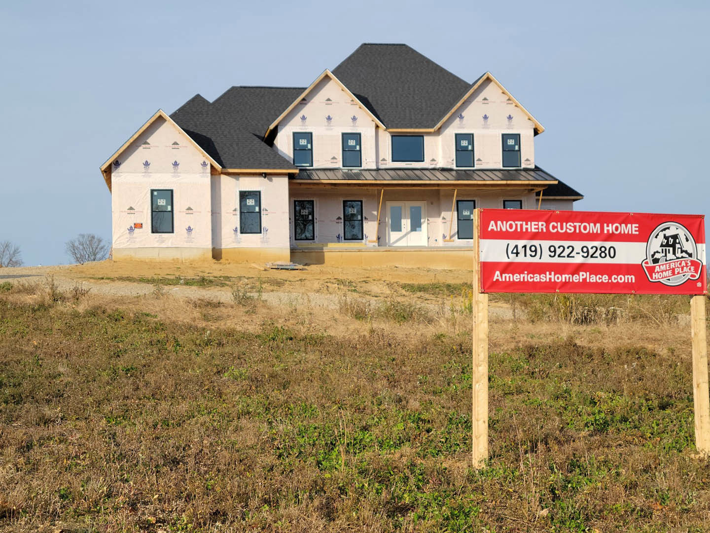 Modern house under construction featuring a black roof, black-framed window, wooden post in grassy field, and red and white banner sign with black text.