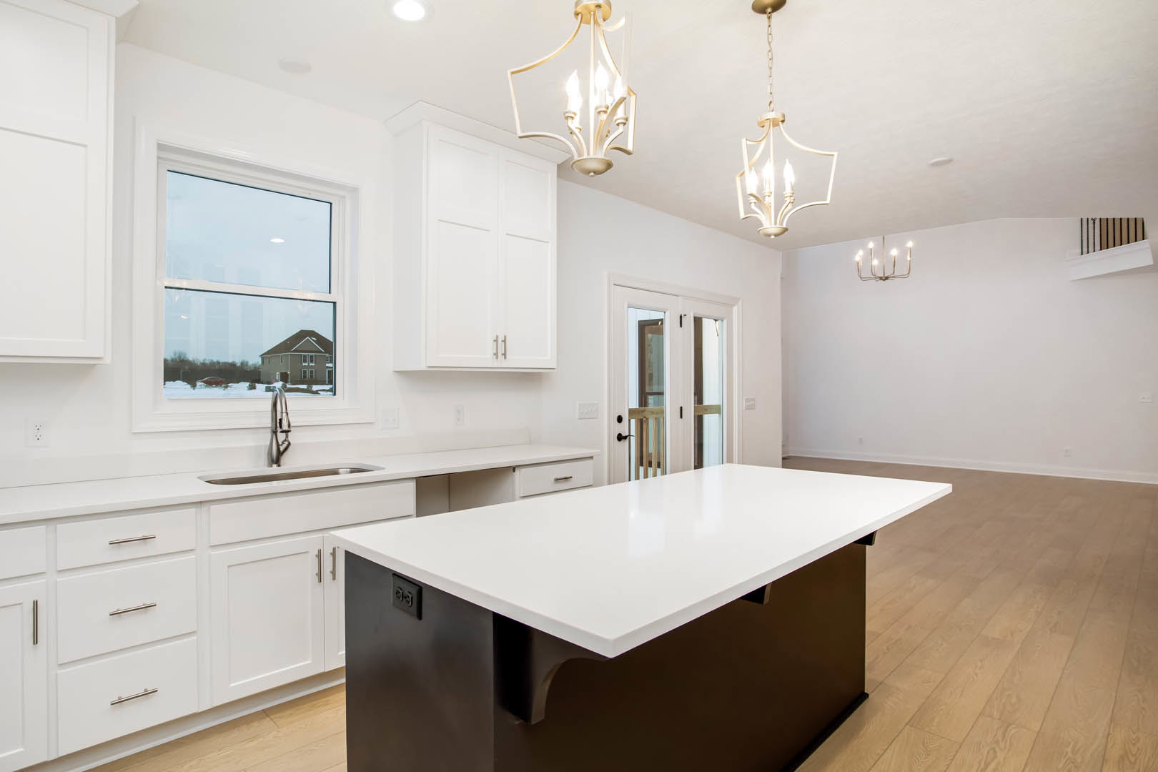 Spacious kitchen featuring a large white island with built-in sink, wood flooring, white cabinetry, glass double doors, modern chandelier, and a window overlooking neighboring