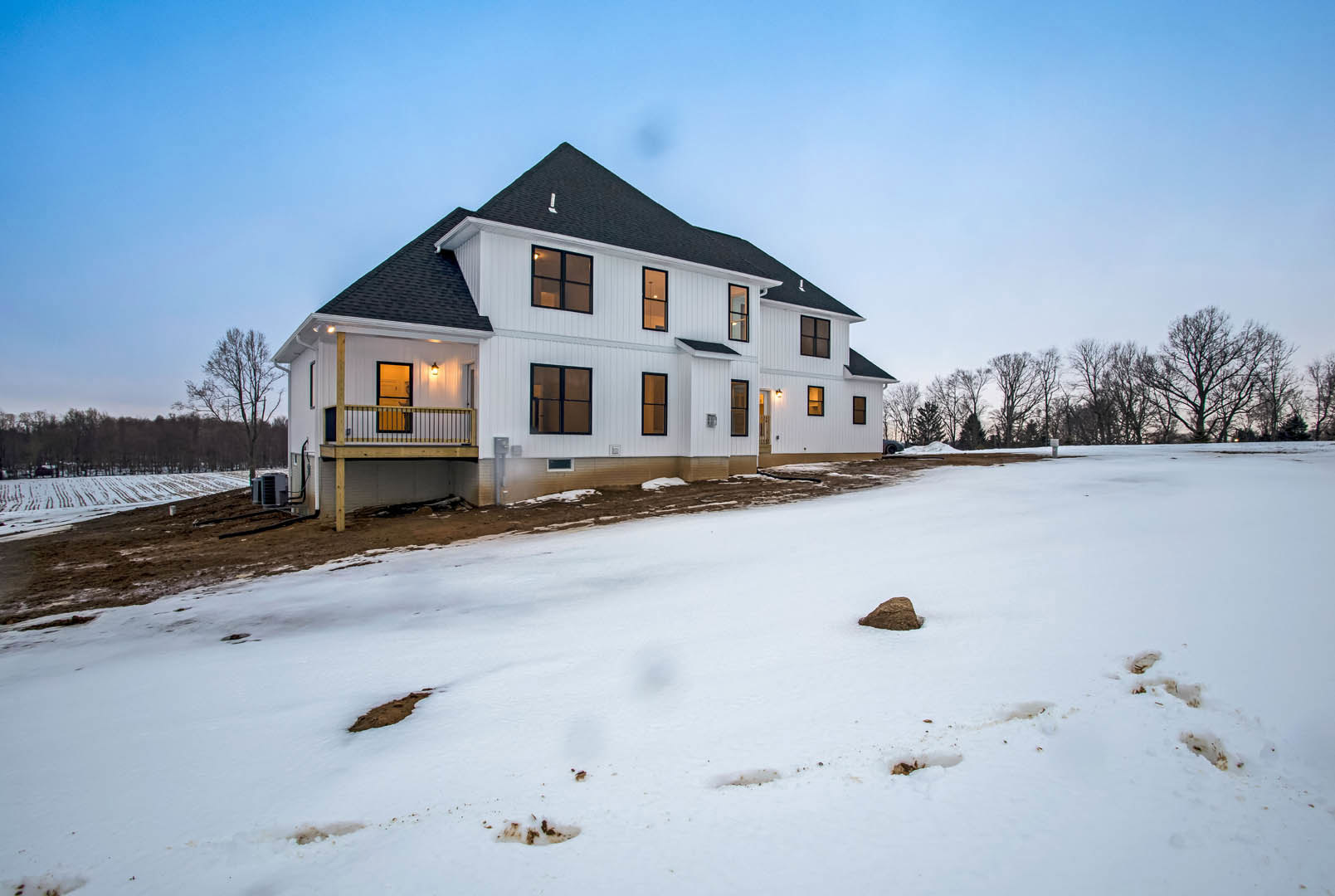 White house with black roof and multiple windows, surrounded by snow-covered ground with visible footprints, bare trees in background, overcast winter sky