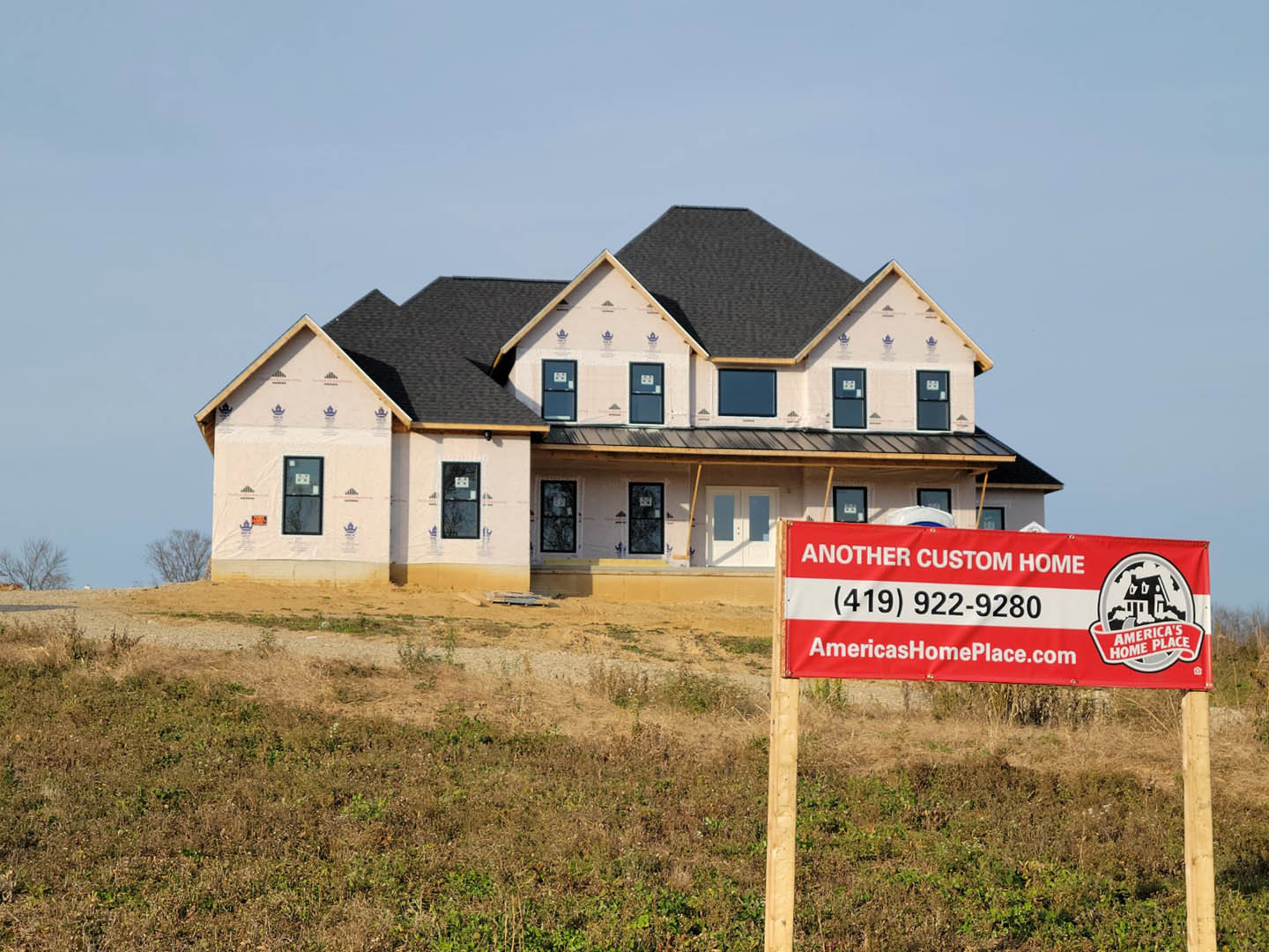 Two-story house under construction with black roof, black-framed window, and red and white sign displaying phone number in grassy front yard