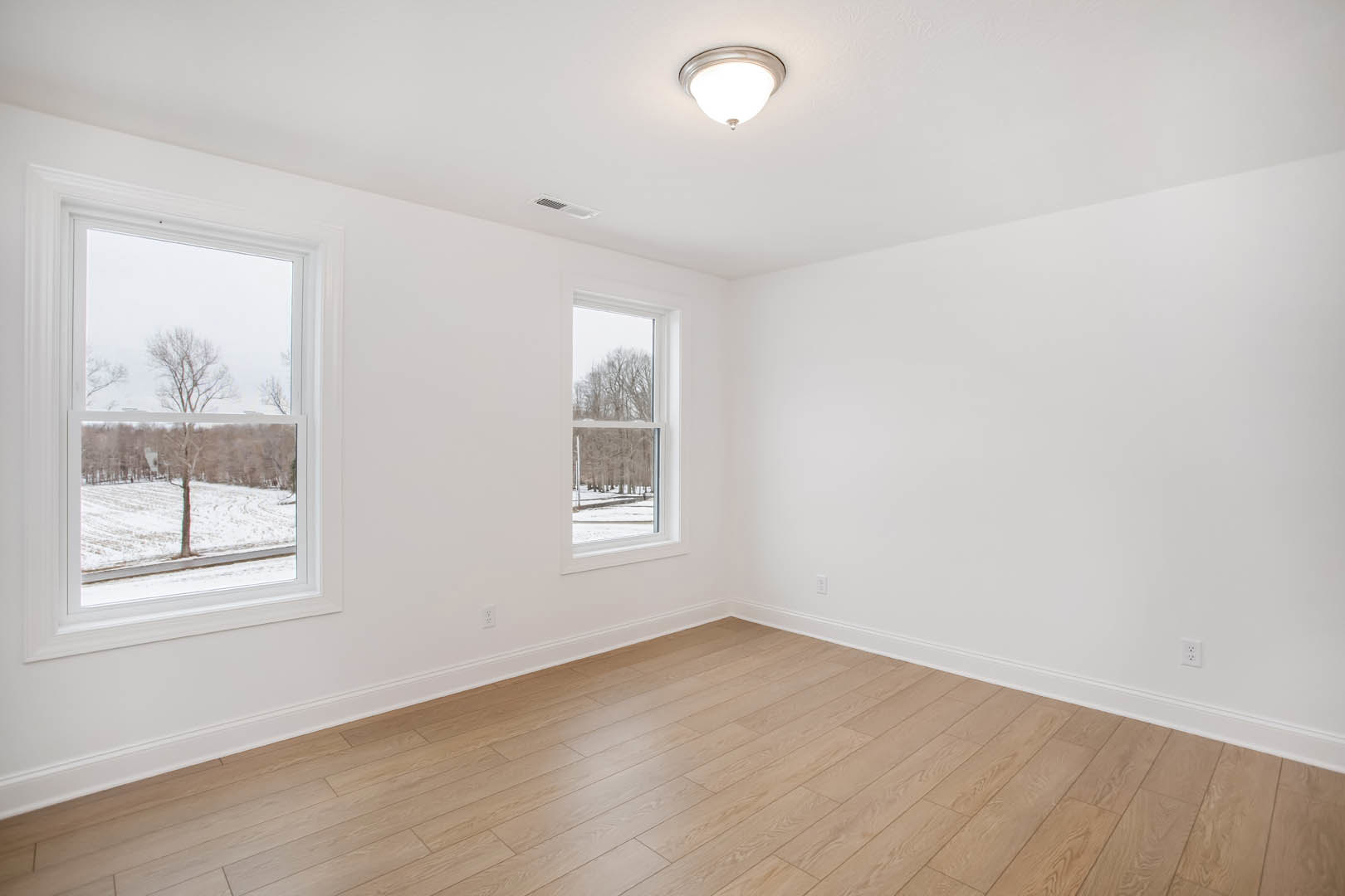 Spacious room with light wood flooring, large windows overlooking snowy field and trees, ceiling light fixture, white plaster walls