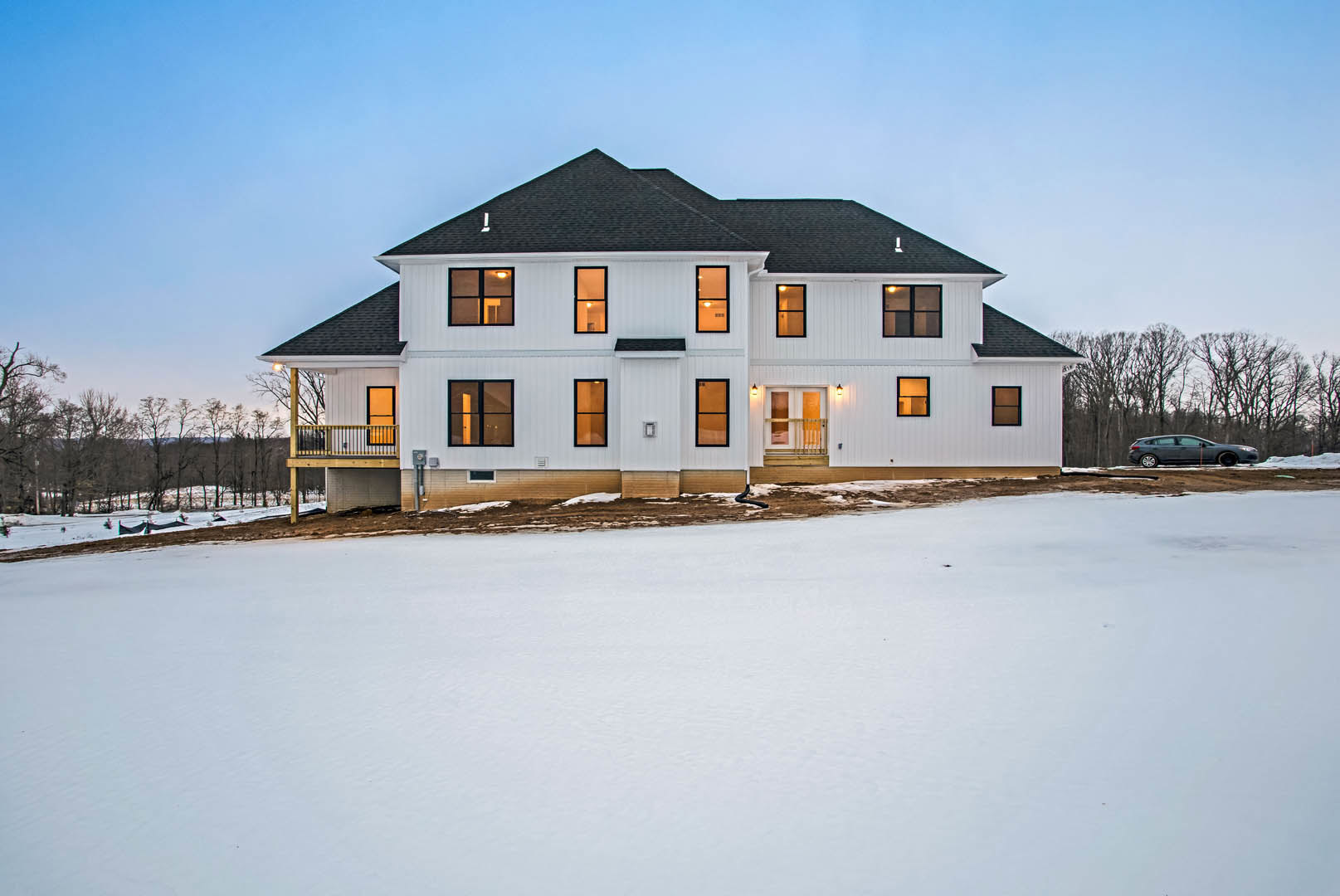 Large white house with black roof and many windows, deck overlooking snowy field, car parked near trees in winter landscape